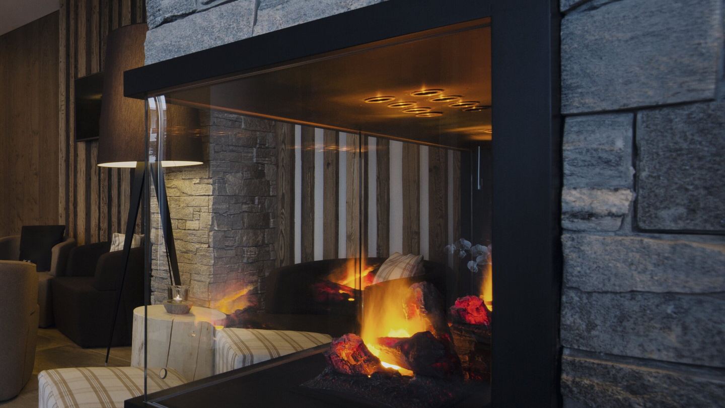 Die Halle: Kamin hinter Glas, Steinwand, Holz und Sessel im warmen Licht
